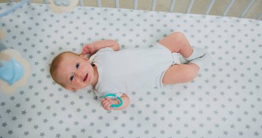 Sırtüstü uzanmış gülümseyen sevimli çocuğun portresi. Kollarını ve bacaklarını bebek karyolasında hareket ettiriyor. Mutlu 2 aylık bebek beşikte diş telleriyle oynuyor, üst manzara.