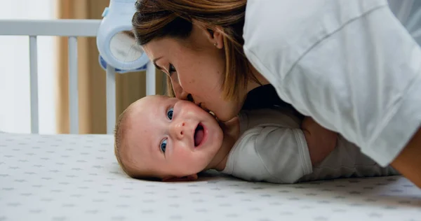 Side view of woman hugging and kissing her toddler as he is lying in white cot while happy smiling looking towards his right side