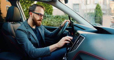 Yakışıklı sakallı, 25-30 yaşlarında modern bir adam. Kendi lüks sınıfı arabasının gidonunun önünde oturuyor ve navigatör kullanıyor..