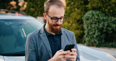 Şehirde beyaz arabanın yanında telefon kullanan şık bir genç. Dışarıda gülümseyen mutlu bir erkek. Teknoloji işadamları çekici modern yaşam tarzı akıllı telefon hırslı cep telefonu..