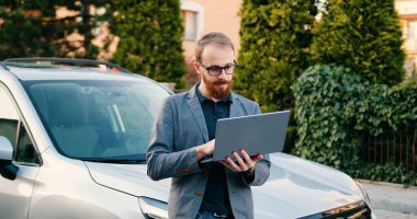 Beyaz, orta yaşlı, güzel bir adam. Siyah arabanın önünde duruyor. Kentte yol kenarında dizüstü bilgisayar kullanıyor..