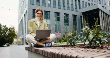 Kendine güveni tam genç bir kadın dışarıda çalışıyor ofis binasının yanında oturuyor laptopla yazı yazıyor kablosuz bilgisayarı ders çalışmak için kullanıyor..