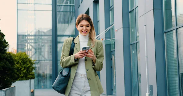 Smiling professional business woman walking while using phone messaging writing texts after work. Girl using smartphone typing text messages walking background modern building.