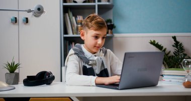 Çocuk çalışmak için dizüstü bilgisayar kullanıyor, online dersler, atölyeler ve yüksek lisans dersleri izliyor. Evdeki modern odada dizüstü bilgisayar derslerine katılan zeki çocuk..