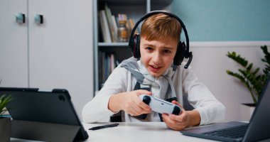 Portrait of excited cute little boy kid sitting on table, emotionally plays on console, loses game, emotions of defeat. Happy child have fun relax after school lessons indoors Game addiction concept.