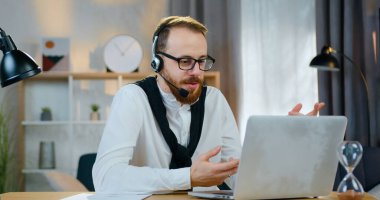 Ev bilgisayarının yanında oturan ve video sohbet yoluyla arkadaşı veya iş arkadaşıyla konuşan kulaklıklı yakışıklı, pozitif pozitif sakallı genç adamın ön görüşü.