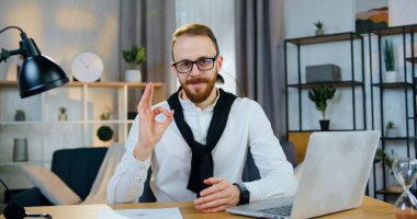 Yakışıklı, kendine güvenen, gözlüklü, kameraya bakan, iyi dekore edilmiş çalışma odasında otururken elinde tabelayla dolaşan, kendine güvenen genç adam.