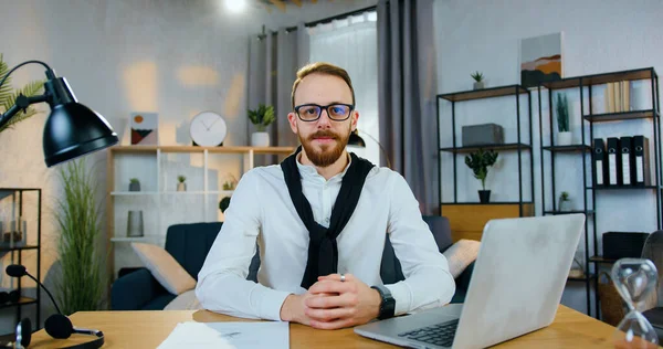 Çekici, kendine güvenen, başarılı, 30 yaşında, sakallı, ofis elbiseli, gözlüklü, iyi dekore edilmiş ev ofisin arka planında kameranın önünde oturan bir ofis müdürü.