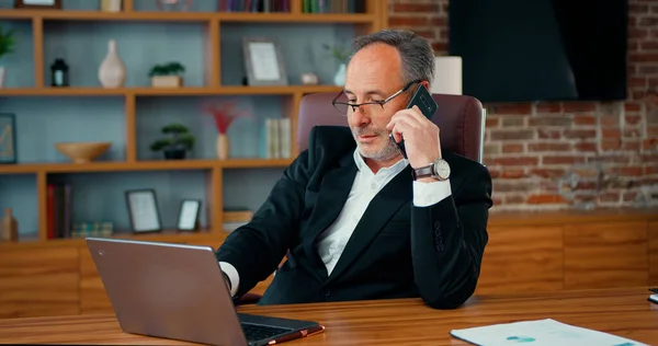 stock image Bearded businessman CEO answering on phone call while using computer. Man office worker manager consulting client by telephone checking information on laptop screen.