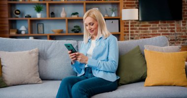 Smiling middle aged lady customer texting mobile sms message playing mobile games using social media apps at home. Millennial woman spending time at home with cell gadget technology.