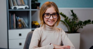 Ev dersinde masasında oturmuş kameraya bakan, gözlüklü, gülümseyen beyaz kız portresi.