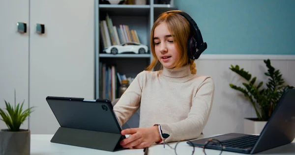 stock image Smart beautiful caucasian 10 years old girl wearing headset watching media on tablet sitting by table in living room at home. Preteeen girl in headphones usin digital tablet and learning English