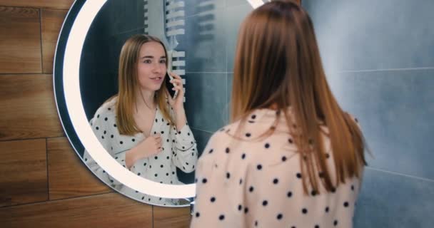 漂亮的20多岁穿着睡衣的年轻女子在家里的前视镜浴室里用智能手机聊天 慢动作 — 图库视频影像