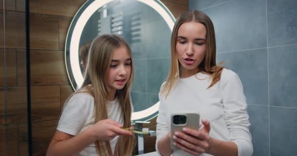 在浴室的早晨卫生程序中 漂亮的年轻女性和小女孩用电动牙刷刷牙 女孩们用智能手机进行社交 慢动作 — 图库视频影像