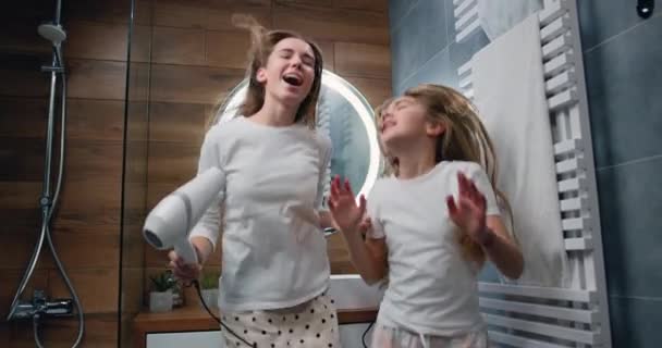 有趣的年轻女人和10岁的女孩 穿着长发睡衣 用吹风机擦干她的长发 一边跳舞 一边在家里的现代浴室里互相望着对方 慢动作 — 图库视频影像