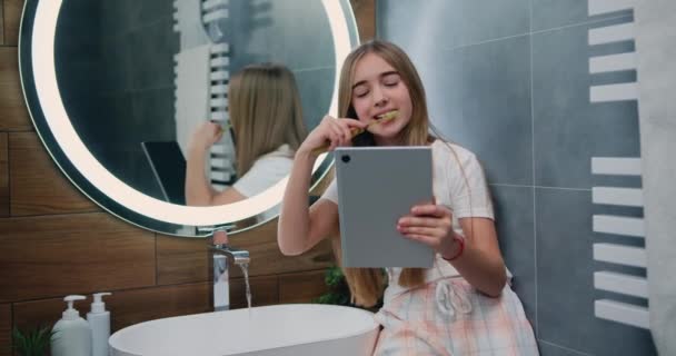 非常有趣的小女孩穿着长发睡衣刷牙 早上在浴室用平板电脑 可爱的女孩在社交网络上使用平板电脑享受酷酷的视频或照片内容 — 图库视频影像
