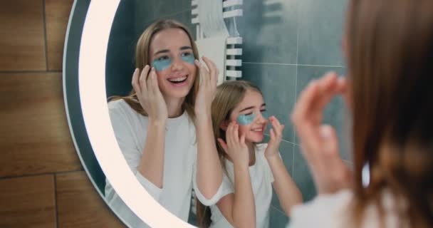 漂亮的女孩和她穿着睡衣的妹妹在家里的浴室里把胶原蛋白贴在眼皮底下 面部护肤的概念 慢动作 — 图库视频影像