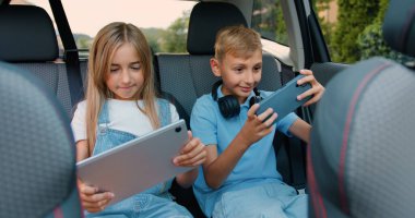 Güzel kız ve erkek çocuklar modern bir arabada otururken telefon ve tabletle oyun oynayarak eğleniyorlar. Küçük kız ve erkek kardeş gülüyor ve mutlu bir şekilde kutlama yapıyorlar.