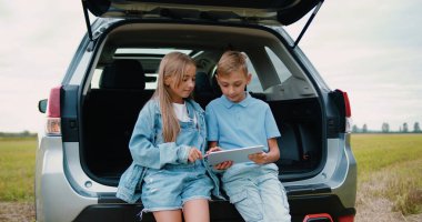 Küçük çocuk ve kız dijital tablette bir şey izliyorlar ve doğada arabanın yanında konuşuyorlar. Yolculuk yapmak, güneşli bir günde arabanın bagajında oturmak..