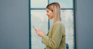 Gülümseyen genç bir kadının caddede yeşil ağaçların yanındaki ofise yürürken cep telefonuna bakışı. Açık havada.