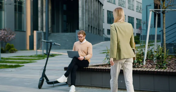 Stock image Portrait of attractive couple of man and woman sitting on bench by modern office building in smart casual business style, working together on laptop, smiling. Outside, freelance worker concept