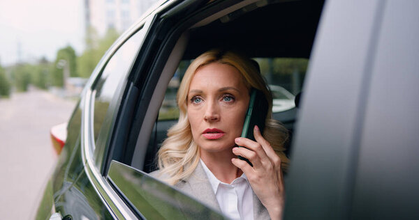 Successful business woman sitting in a luxury white car talking on the phone view from the street through the window. Lady manager in a suit. Communicates with partners. Business conversation.