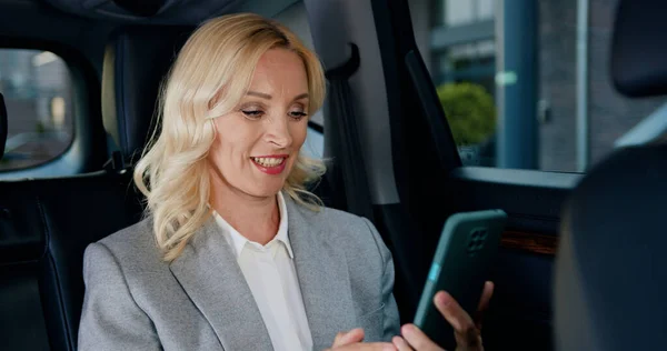 Portrait middle age woman in suit talking by video call on mobile phone sitting in the car. Businesswoman having video chat with smartphone at remote workplace.