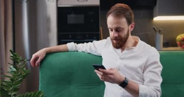 Yakışıklı beyaz adam mutfakta kanepede oturuyor ve akıllı telefon kullanarak sosyal medyada geziniyor, video izliyor. İnsanlar ve modern teknoloji kavramı. Yavaş çekim.