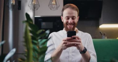 Mutfaktaki koltukta dinlenirken cep telefonu kullanan mutlu beyaz adam. Üç gün sakallı bir erkek gülümseyerek başını zevkle sallıyor. Yavaş çekim.