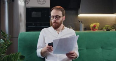 Mutfaktaki kanepede oturan hesap makinesini kullanarak mali işlerle ilgilenen bir adam. Finansal konsept. Evde verileri analiz eden odaklanmış bir adam. Yavaş çekim.