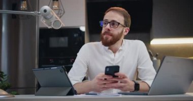 Masada oturan neşeli iş adamı modern bir mutfakta gülümseyen bir akıllı telefon kullanıyor. Serbest çalışan erkek elinde telefonla mesaj teknolojisi uygulaması tutuyor, evde e-posta yazıyor. Yavaş çekim.