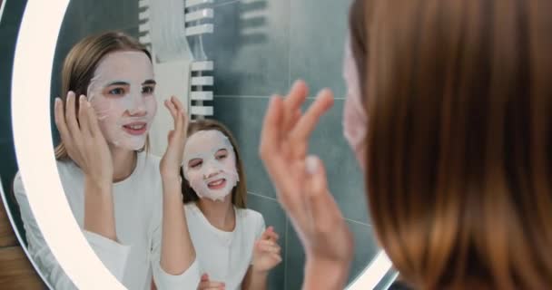 两个漂亮的女孩站在家里的浴室里 面罩着化妆品 看着镜子 皮肤护理和美容美发概念 慢动作 — 图库视频影像