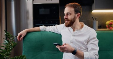 Yakışıklı beyaz adam mutfakta kanepede oturuyor ve akıllı telefon kullanarak sosyal medyada geziniyor, video izliyor. İnsanlar ve modern teknoloji kavramı.