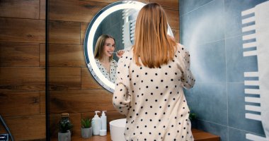 Güzel görünümlü beyaz bir kadın aynaya bakıyor ve banyoda dişlerini fırçalıyor. Sağlıklı dişler, günlük hijyen kavramı.