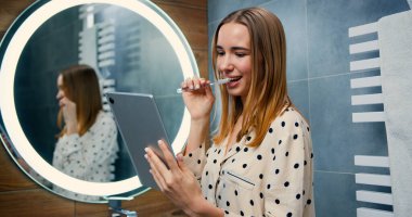 Uzun saçlı, pijamalı, dişlerini fırçalayan ve sabah evde tablet aletine bakan beyaz bir kadın.