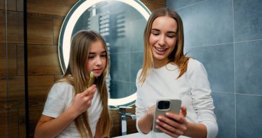 Güzel genç bir kadın ve küçük bir kız sabah banyoda diş fırçasıyla diş fırçalarken, kızlar sosyal ağ için akıllı telefon kullanıyor.