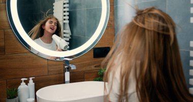 Güzel küçük kız saçını saç kurutma makinesiyle kurutur, banyoda aynanın önünde dikilirken şarkı söyler ve gülümser. Saç kurutma makinesiyle uzun ıslak saçlarını kurutan tatlı bir kız.