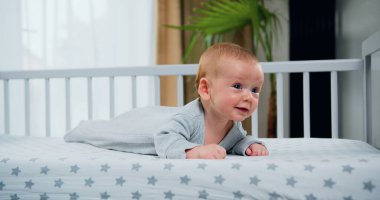 Güzel mavi gözlü, gülümseyen bebek portresi. Üç aylık erkek bebek başı yukarıda yüzüstü yatar. Beyaz yatakta sürünen tatlı bebek, mutluluk ve eğlence..