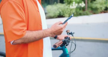 Modern şehir binasının arka planında bisiklete yaslanırken akıllı telefon kullanarak mesaj yazan yakın plan adam. Yavaş çekim.