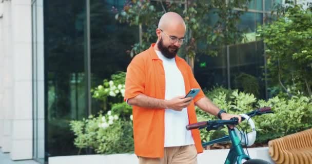 都会を自転車で歩きながらスマホを使いカジュアルな服装や眼鏡をした白人髭の男 通勤は市内で持続可能なライフスタイルを働く スローモーション — ストック動画