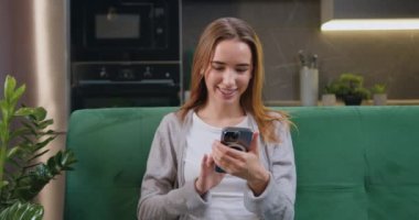 Mutfak arka planında yeşil bir koltukta otururken cep telefonu kullanan mutlu beyaz kadın. Sarı saçlı, gülümseyen kız rahat koltukta dinleniyor ve akıllı telefondan internete takılıyor. Yavaş