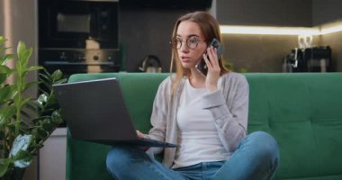 Telefonda dizüstü bilgisayarla konuşan sevimli genç bir kadın mutfak arka planında oturuyor, mutlu bayan müşteri internet sitesinden alışveriş siparişi konseptini doğrulamak için mobil arama yapıyor. Yavaş çekim