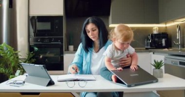 Anne, küçük çocukları oynarken ve onu rahatsız ederken dizüstü bilgisayarla çalışmaya çalışıyor. Uzaktan çalışma ve evde çocuklarla eğlenme. Serbest çalışıyorum, oğlumla evde çalışıyorum. Yavaş çekim.