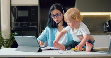 Genç anne ve iş CEO 'su bebek oyun oynarken ve dikkati dağınıkken mutfak masasında dizüstü bilgisayarıyla çalışıyor. Annelik ve çalışma aynı anda. Yavaş çekim.