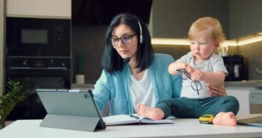 Kulaklıklı bir kadın dizüstü bilgisayar kullanıyor ve evde online konferans yapıyor. Anne ve küçük oğlu mutfaktaki çalışma masasında oynuyorlar ve çalışıyorlar. Annemi serbest çalışan olarak kabul et. Yavaş çekim