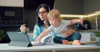 Anne, erkek bebekle uzaktan çalışıyor. Uzak iş konsepti. Kadın evde, mutfakta çalışmaya çalışıyor, bebek çocuk masasında oturuyor. Yavaş çekim.