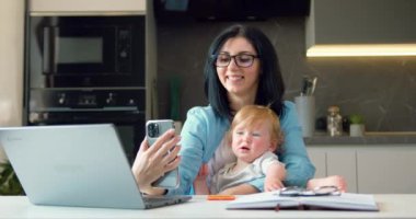 Anne, küçük oğluyla akıllı telefonda özçekim yapıyor, mutfakta kameraya poz veriyor ve gülümsüyor. Aşk, aile bağı ve takım çalışması. Yavaş çekim.