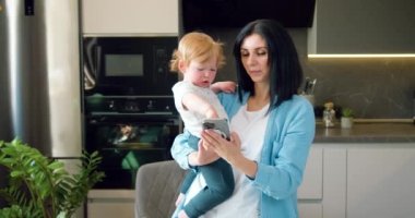 Kadın ve küçük oğlu telefona bakıyor ve modern bir mutfakta gülümsüyorlar. Anne evde çalışırken çocuğuyla oynar. Aşk, aile bağı ve takım çalışması. Yavaş çekim.