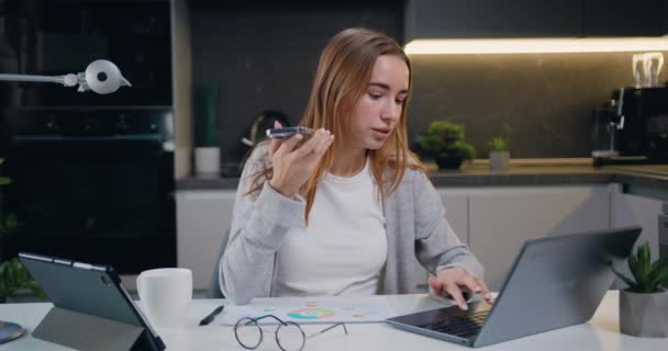 专注的年轻高加索女人 坐在办公桌前办公 用手提电脑在手机上记录音频讯息 千年女性使用笔记本电脑 并在智能手机上记录虚拟数字语音 — 图库视频影像