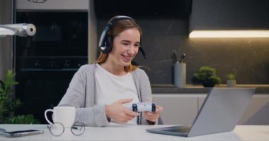 Güzel görünümlü, heyecanlı, kulaklıklı, video oyununda zaferi kutlayan kulaklıklı, neşeli, genç, beyaz bir kız. Yumruklarını sıkarak ve gülümseyerek, boş zamanlarını oturma odasında geçiriyor. Yavaş çekim